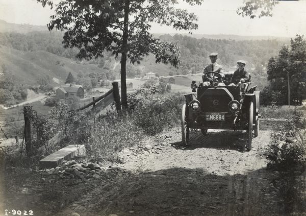 Man and boy (possibly father and son) driving an International Model M truck on a rough dirt road overlooking farmland in a valley.