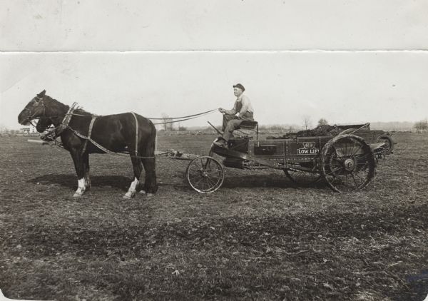 Side view of a man posing in a field on a horse-drawn International Low-Lift manure spreader.