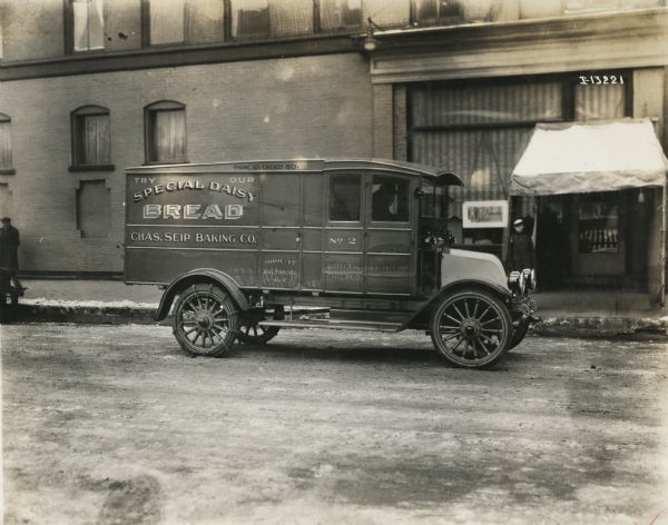 View from across the street of a man driving an International Model M truck for the Chas. Seip Baking Company on a snow-covered street. Tire chains have been placed on the truck's rear wheels. A doorman is standing on the sidewalk near an entrance to a building. Text on the side of the truck advertises "Special Daisy" bread.