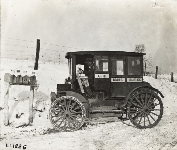 Mail man driving an International Model M U.S. Mail truck that was part of the Rural Free Delivery service. The truck was equipped with tire chains to drive through snow and is parked next to several mailboxes.