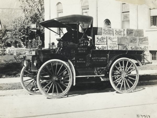 Man in the driver's seat of an International Model M truck hauling crates of beer labeled "Grand Rapids Brewing Company" and "Petersen Brewing Company." The truck is parked on a road outside a large building. Also with him in the open cab is a dog.