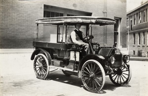 Man seated in a parked International Model E truck near brick buildings. The truck has a covering that can be raised and lowered over the bed and cab, and appears to have a telephone company logo on its door.