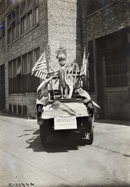 International Model E truck adorned with American flags, International Harvester pennants and a display of "Prosperity" or "Prospy" for short. "Prospy", the small statue atop the truck, has a body of a corn cob and a shield of the IH logo. A sign on the front of the truck reads: "Prosperity, Prospy for Short."