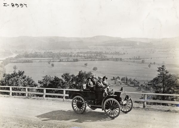 Slightly elevated view of a man driving three women in an International Model M truck along a rural road. In the background is a valley with farm buildings, a river and a bridge.