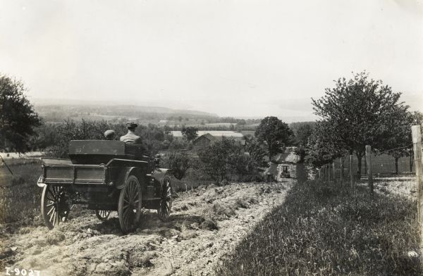Man and boy, possibly father and son, drive an International Model M  truck on rough dirt road near a fence.