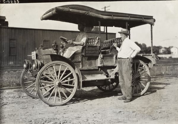 Two men with an International Model M truck. The truck is parked in a dirt lot near railroad tracks. Lettering on the truck identifies the "Barrington Mercantile Company."