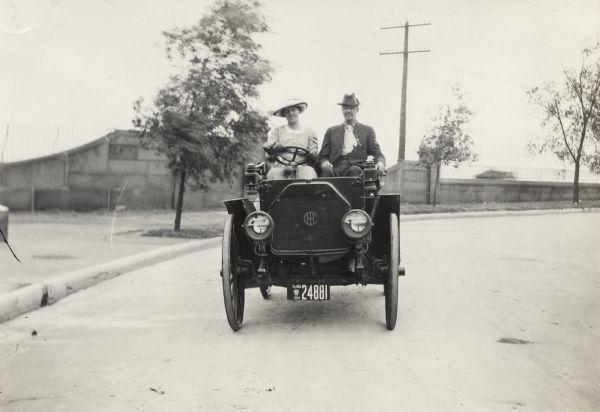 Woman driving an International Model M truck with man in the passenger seat. The two are driving down a road, possibly overlooking a body of water.