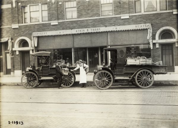 Two International trucks parked outside Atkin & Tharp grocery store with shipments of produce. The grocer is leaning against one of the trucks and the delivery man is holding a crate of produce. A man is sitting in the driver's seat of the truck on the right. One of the trucks has a sign with lettering identifying the truck: "W. Terpstra & Son, Gardeners".