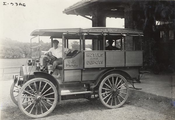 Young man sitting in an International Model M truck used for transportation of passengers, the U.S. Mail, and the transport of luggage to hotels and depots. The truck, or bus, is parked near a brick building, possibly a depot.