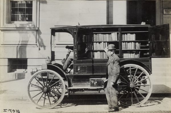 Man in a bowler hat standing next to an International Model M truck used as a bookmobile for the Washington County Library. The shelves of books are accessible through double doors on the side of the truck. Another man is sitting in the driver's seat. A sign near the door of the library appears to read: "Washington County Free Library."
