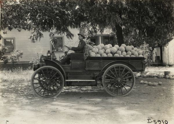 Man parked in an International Model M truck outside a suburban or rural home with an overflowing bed of squash or pumpkins.