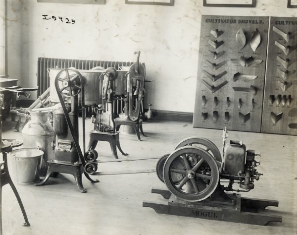 A Mogul stationary engine belted to one of three cream separators on display in dealership showroom. Large display boards in background show various cultivator shovels.