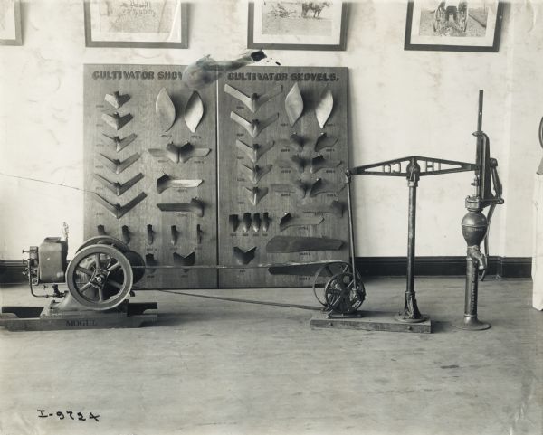 A Mogul stationary engine is belted to walking beam pump jack for display in a dealership showroom. Two large display boards in background show the various cultivator shovels offered by the dealer.