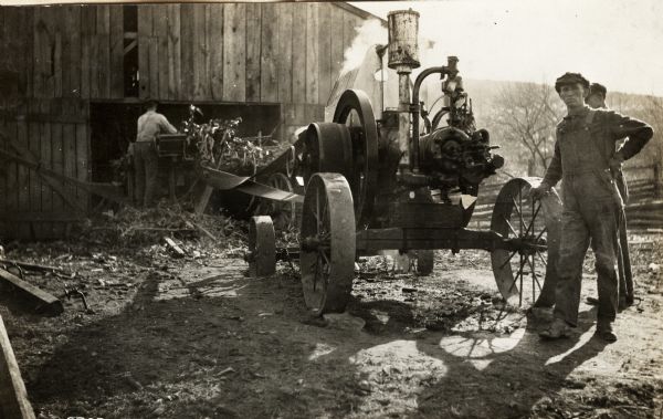 Men standing in a farmyard with a Mogul engine running what appears to be a belt-driven ensilage cutter, which is near the open door of a barn.