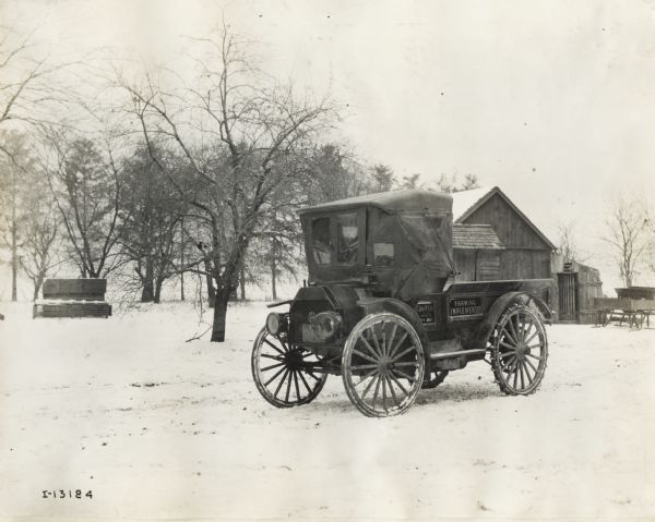 International Model M truck parked in snow on rural land. The truck is used by John W. Esch, a farm implement dealer (likely for International Harvester). The truck is equipped for the winter weather with tire chains and a cover for the cab.
