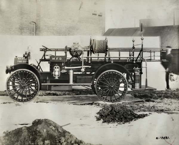 International Model E 1916 fire truck operated by the Marion Fire Department. Left side view of truck displays various equipment used for fire extinction. A man is standing near the rear of the truck on the right and holding a sheet for a backdrop. Brick buildings are in the background.