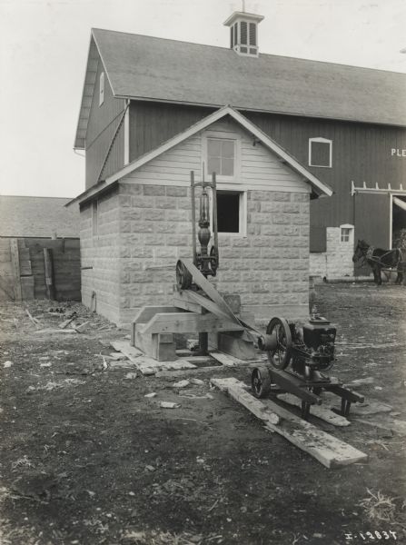 Mogul Jr. 1 HP engine attached with a belt to a water pump. Water flows into a nearby small stone building on a farm. A barn is in the background, as well as two work horses.