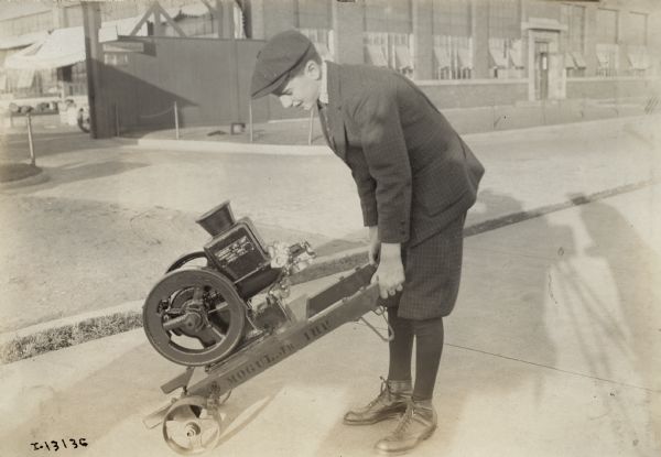 Young man outdoors holding a Mogul Jr. 1 HP engine mounted on a skid with two wheels. An industrial building or a factory is in the background. The shadow of a photographer and camera is also visible.