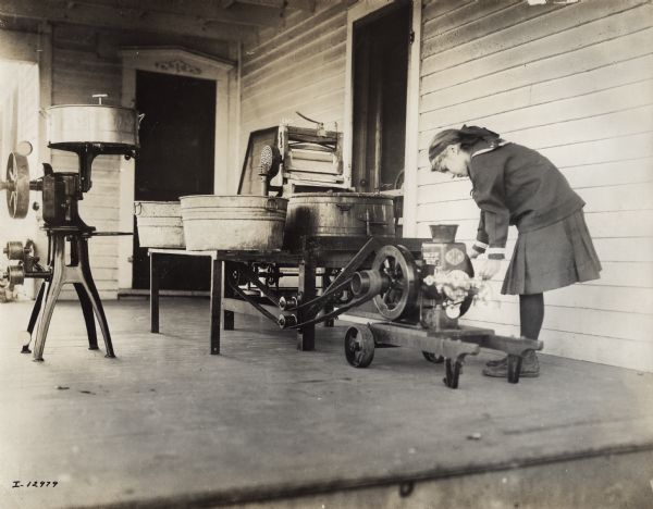 A small girl using a Mogul Junior 1 HP stationary engine to power a washing machine located on the porch of a house. There is a cream separator on the left.
