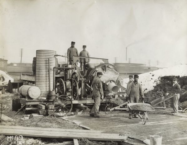 Men at a construction site using an International stationary engine to power a concrete mixer. Two of the men have wheelbarrows filled with concrete.