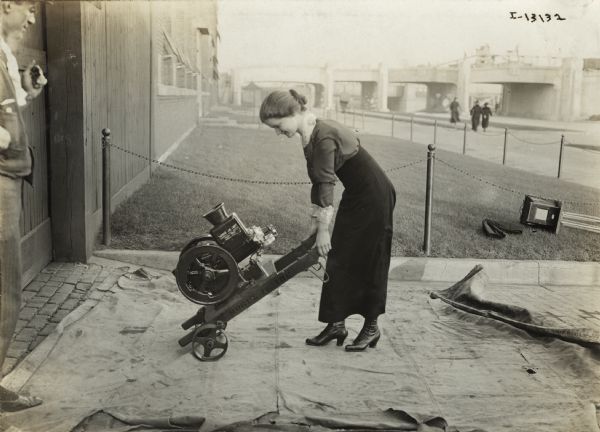 A young woman outside industrial building or factory holds a Mogul Jr. 1 HP engine mounted on a skid with two wheels. A man stands nearby on the left. On the lawn in the background is a box camera on a tripod lying on the ground.