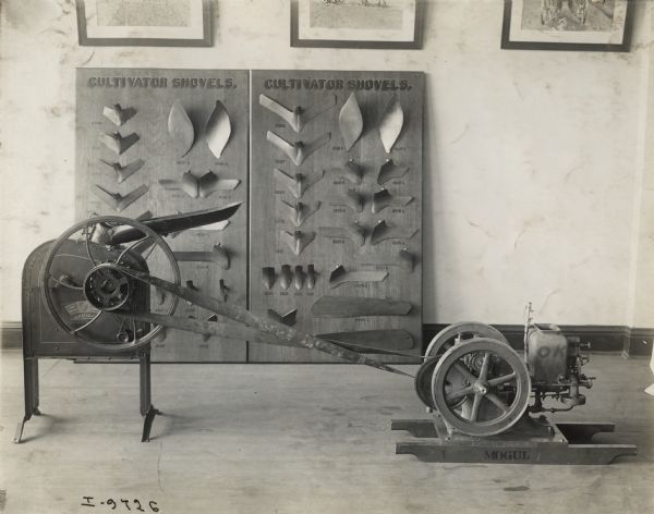 Mogul engine belted to a corn sheller in the showroom of an International Harvester dealership. Two display boards of cultivator shovels are propped against a wall in the background.