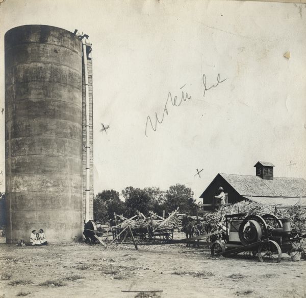 Men using a stationary engine to power a silo filler. Two men are loading the silo filler while another man is standing in a horse-drawn wagon. Another man is standing at the top of the silo. Two women and small girl along with a dog are sitting on the ground beside the silo.