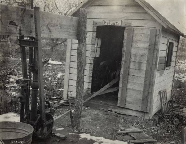A stationary engine (possibly a Mogul) in a small wooden shed is used to power a belt-driven water pump positioned nearby. A small wood sign reads 'private' over the top of the shed door.