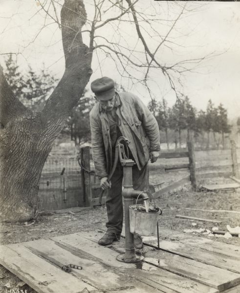 A man stands on a wood platform and uses a hand operated water pump to fill a metal bucket.
