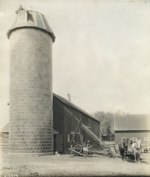 A farmer with a team of two horses working with an elevator or silo filler near farm buildings and a silo. A Titan stationary engine is powering the elevator, which was made by the Sandwich Manufacturing Company of Sandwich, Illinois. Underneath the wagon a dog is resting, and on top of a windmill seen above the barn roof, there appears to be a man standing on a platform.