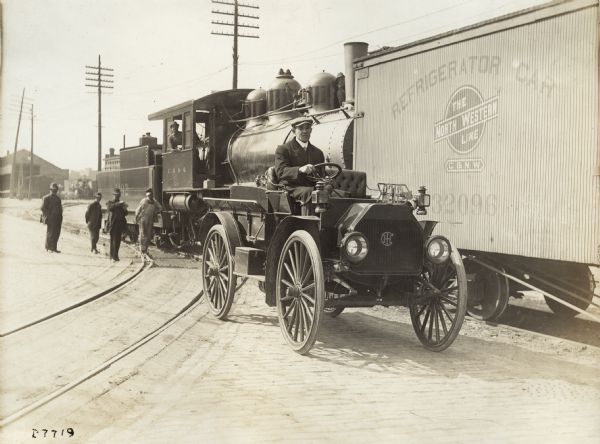 A man in an International Model M truck parked in a railroad yard beside a freight train. In the background is a locomotive and a refrigerator car. The train is operated by North Western Line. A small group of men stand behind the motor truck, and the engineer is leaning out of the window of the locomotive.
