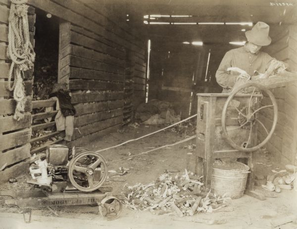 A man in open doorway of a farm building using a Mogul Jr. 1 H.P. stationary engine to power a belt-driven Keystone corn sheller.