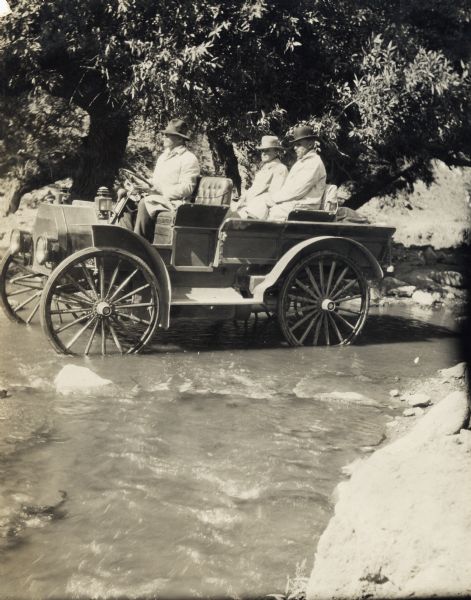 Three men in an International Model M truck driving through a stream in a wooded area.