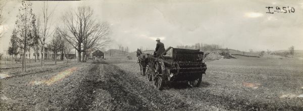 Panoramic view of man operating a horse-drawn manure spreader on farmland close to farmhouse and farm buildings.