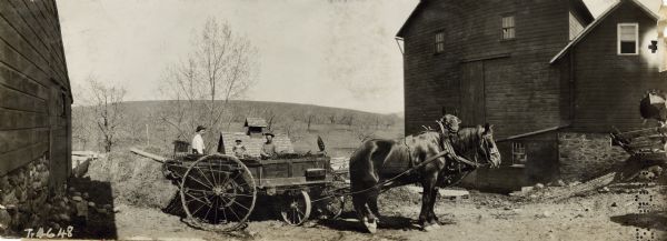 Panoramic view of group of three men standing behind a partially loaded horse-drawn Kemp No. 2 20th Century manure spreader between farm buildings.