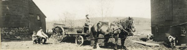 Panoramic view of man on horse-drawn Kemp No. 2 20th Century manure spreader. Sitting on the ground on the left is a man and young boy (possibly father and son). Cattle are standing near a barn on the right.