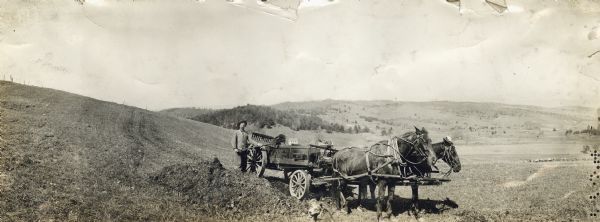 Panoramic view of farmer with dog loading manure onto horse-drawn Kemp No. 1 20th Century manure spreader at the base of a hill on farmland.