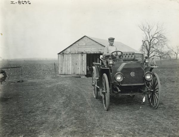 Man driving an International Model M truck on farmland. In the background is a shed near a field.