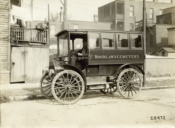 Man parked on urban side street in an International Model M truck operated by Woodlawn Cemetery (possibly as a hearse).