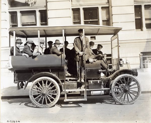 Group of men seated in an International Model E motor truck parked outside large stone building. One man stands inside truck near its side steps. The truck appears to be in use as a bus.