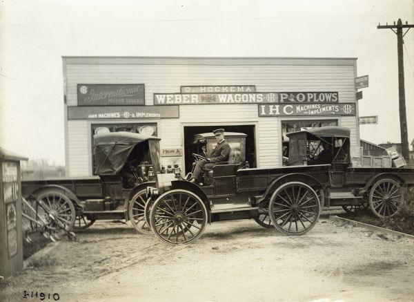 Men sit in four International trucks outside of an International Harvester dealership. One of the trucks is a Model M operated by Terpstra & Son Gardeners. Signs on the dealership advertise Weber wagons, P&O plows and International motor trucks. A name over the front door reads: "O. Hockema".