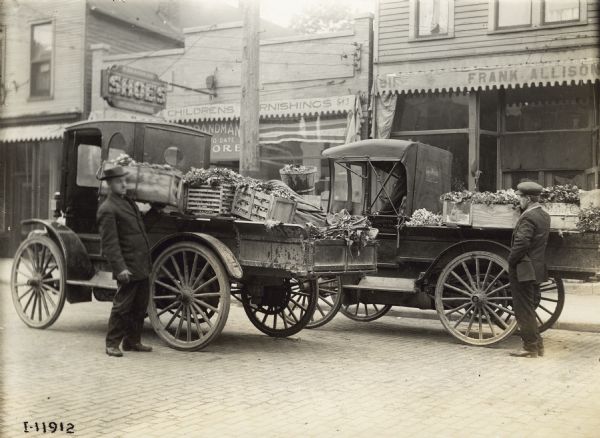 Two men with trucks loaded with produce outside the storefront of Frank Allison. One of the trucks is an International Model M operated by Terpstra & Son Gardeners.