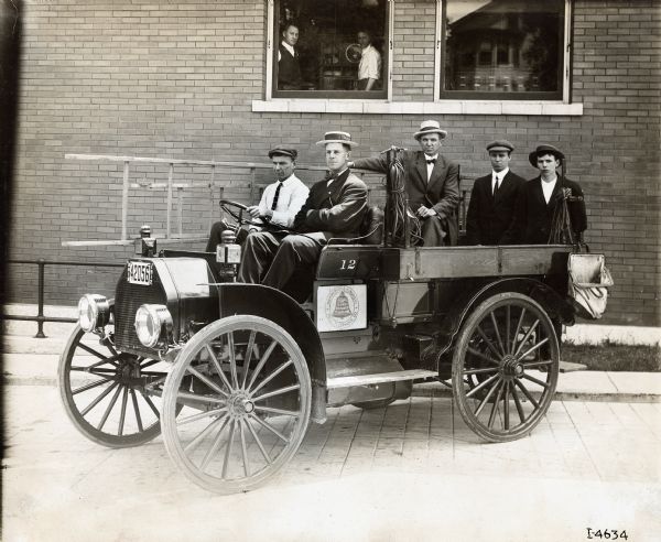 Several men posing in an International Model M truck used by the Cincinnati & Suburban Bell Telephone Company. The truck is parked outside a brick building with large windows, from which two men can be seen looking out from an office.