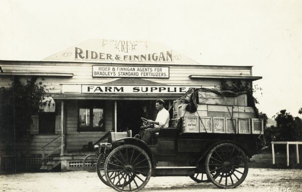 Man parked in an International Model M truck loaded with freight outside the "Rider and Finnigan" storefront. Rider and Finnigan was a farm supply dealer, and were agents for "Bradley's standard fertilizer."