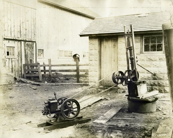 Water pump hooked up to a Mogul stationary engine outside a small stone farm building. Water flows from pump into building through small piping running from the pump through a wall.