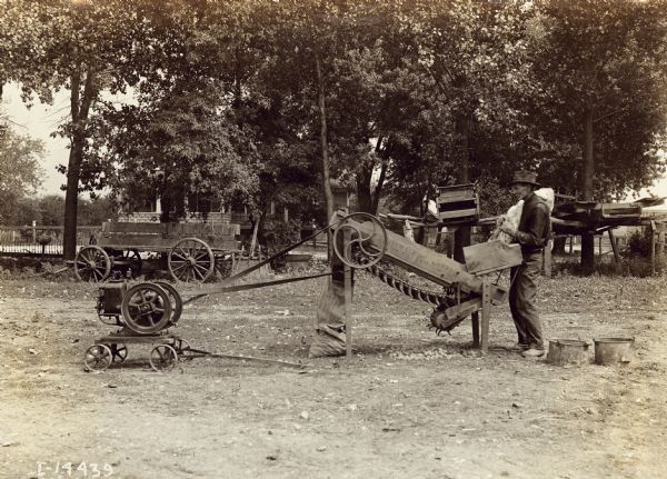 A man in a yard is using a Mogul stationary engine to power farm machinery, possibly a feed grinder. In the background is a house behind trees.
