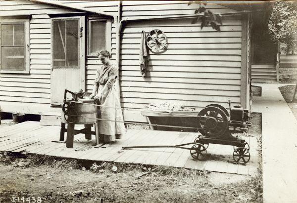 A woman standing on the back porch of a home uses a Mogul stationary engine to power a washing machine.