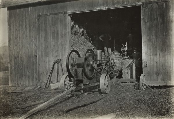 Men standing inside a barn and use an International Motor Baling Press, or hay press to bale hay.