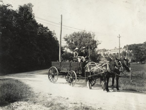 A man and boy seated in a loaded Sterling wagon pulled by a team of horses on a dirt road. In the far background stand several buildings, including residences.