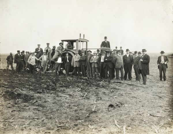View from distance of a group of approximately 40 well-dressed men with Titan tractor on farmland.